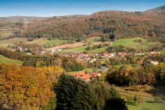 Blick über den Ortsteil Bockenrod Panoramablick von oben auf ein idyllisches Dorf, eingebettet in ein Tal einer sanften Hügellandschaft. Die umliegenden Wälder leuchten in intensiven Herbstfarben von Gelb, Orange und Rot, durchsetzt mit grünen Feldern. Am Horizont ist ein klarer blauer Himmel zu sehen.