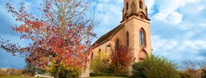 Christuskirche im Krautweg Eine Kirche aus hellem Sandstein mit einem hohen, spitzen Kirchturm und einem Kreuz auf der Spitze steht auf einem grünen Hügel. Links im Vordergrund steht ein Baum mit leuchtend roten Herbstblättern, der die herbstliche Atmosphäre betont. Der Himmel ist strahlend blau mit weißen Wolken.