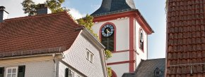 Evangelische Kirche in Reichelsheim Ein hoher Kirchturm mit einem dunklen, spitz zulaufenden Schieferdach und einem weißen, rot umrandeten Glockenturm dominiert das Bildzentrum. Er zeigt blaue Zifferblätter. Davor stehen traditionelle Häuser: links ein weißes Haus mit grünen Fensterläden und rotem Ziegeldach, rechts ein weiteres Haus mit rotem Dach, das nur teilweise sichtbar ist. Ein gepflasterter Weg führt zwischen den Gebäuden hindurch. Der Himmel ist strahlend blau mit einigen weißen Wolken, und die Szene wird von warmem Sonnenlicht beleuchtet.
