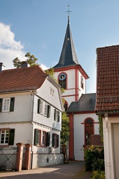 Ein hoher Kirchturm mit einem dunklen, spitz zulaufenden Schieferdach und einem weißen, rot umrandeten Glockenturm dominiert das Bildzentrum. Er zeigt blaue Zifferblätter. Davor stehen traditionelle Häuser: links ein weißes Haus mit grünen Fensterläden und rotem Ziegeldach, rechts ein weiteres Haus mit rotem Dach, das nur teilweise sichtbar ist. Ein gepflasterter Weg führt zwischen den Gebäuden hindurch. Der Himmel ist strahlend blau mit einigen weißen Wolken, und die Szene wird von warmem Sonnenlicht beleuchtet.