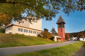 Johanneskirche im Ortsteil Beerfurth Ein modernes, weißes Kirchengebäude mit einer Reihe von Fenstern, das auf einem grünen Hang liegt. Daneben steht ein markanter, rot-oranger Glockenturm mit einem spitzen, dunklen Dach und offenen Lamellen im oberen Bereich. Eine Straße führt am Gebäude vorbei, und im Hintergrund sind weitere Häuser eines Dorfes sowie bewaldete Hügel in herbstlichen Farben unter einem klaren, blauen Himmel zu sehen. Grüne Blätter eines Baumes rahmen die Szene von oben ein und deuten auf einen sonnigen Herbsttag hin.