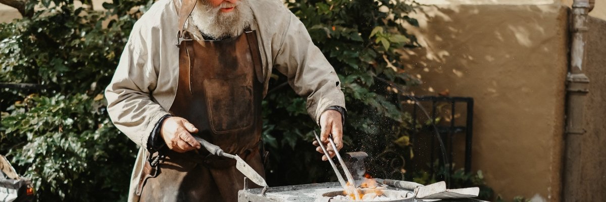 Älterer Mann in mittelalterlichen Klamotten arbeitet an einem Amboss