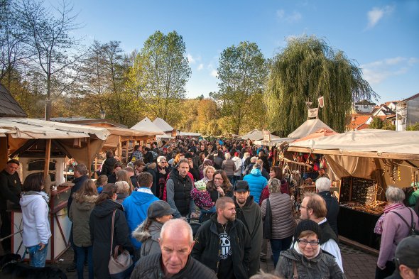 Ein belebter Freiluft-Markt, möglicherweise ein Mittelaltermarkt, an einem sonnigen Tag. Zahlreiche Menschen unterschiedlichen Alters schlendern zwischen rustikalen Marktständen mit Holzdächern und hellen Stoffplanen umher. Die Stände bieten verschiedene Waren an, darunter Schmuck und handwerkliche Produkte. Im Hintergrund sind grüne und vereinzelt herbstlich gefärbte Bäume unter einem strahlend blauen Himmel mit wenigen Wolken zu sehen. Am rechten Bildrand sind einige Hausdächer erkennbar. Die Szene ist von einer lebhaften, heiteren Stimmung geprägt.