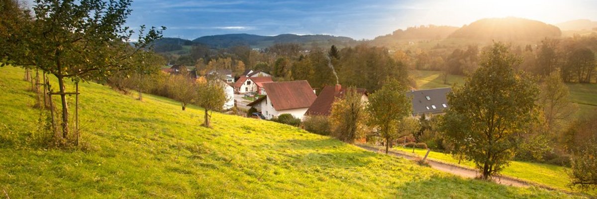 Ober-Gersprenz Ein sonniger Herbsttag zeigt einen steilen grünen Wiesenhang im Vordergrund, mit jungen Bäumen auf der linken Seite. Dahinter liegt ein kleines Dorf mit Häusern, die rote Dächer und helle Fassaden haben, umgeben von weiteren Bäumen und bewaldeten Hügeln. Die tief stehende Sonne taucht die rechte Seite der Landschaft in ein warmes goldenes Licht unter einem blauen Himmel.