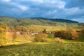 Ober-Kainsbach Weitläufige ländliche Herbstlandschaft mit einem sonnenbeschienenen grünen Hang im Vordergrund, auf dem Bäume mit gelb-goldenem Laub stehen. Dahinter erstreckt sich ein weites Tal mit grünen Feldern und mehreren kleinen Siedlungen aus hellen Häusern mit roten Dächern. Am Horizont erheben sich dunkelgrüne, bewaldete Hügel. Der Himmel ist dramatisch mit dunklen Wolken, durch die das Sonnenlicht bricht und bestimmte Bereiche der Landschaft warm beleuchtet.
