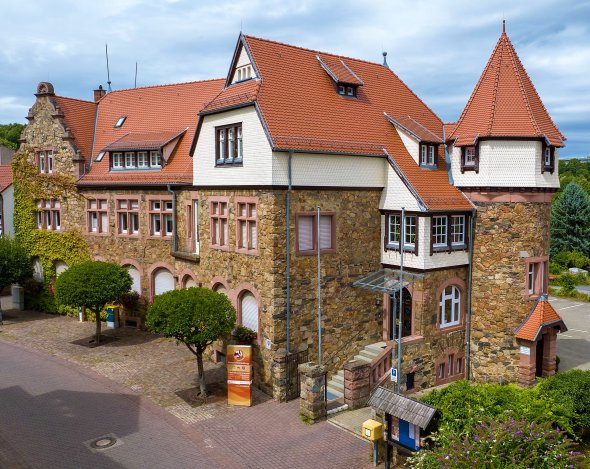 Ein beeindruckendes, historisch wirkendes Gebäude mit Natursteinmauern in warmen Erdtönen und einem markanten roten Ziegeldach. Das Gebäude weist mehrere Giebel, Gaubenfenster und einen auffälligen runden Turm mit spitzem, konischem Dach auf der rechten Seite auf. Einige Giebelflächen und der obere Teil des Turms sind mit weißer Schindelverkleidung versehen. Vor dem Gebäude befindet sich ein gepflasterter Vorplatz mit zwei kleinen, gepflegten Bäumen. Ein orange-gelbes Banner mit der Aufschrift "Gemeindeverwaltung" und "Touristik-Information" steht links, und rechts ein hölzernes Informationsschild mit Postkasten. Die Fassade auf der linken Seite ist teilweise mit Efeu bewachsen. Der Himmel ist leicht bewölkt.