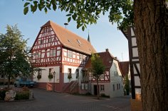 Eine idyllische Straßenszene an einem sonnigen Tag. Im Zentrum steht ein großes, zweistöckiges Fachwerkhaus mit markanten roten Holzbalken und weißen Wänden. Auf dem steilen, roten Ziegeldach ist deutlich eine Storchennestplattform zu erkennen. Hinter dem Fachwerkhaus ragt die Spitze eines Kirchturms in den klaren blauen Himmel. Links im Bild ist ein kleiner, blauer Lastwagen auf einem gepflasterten Platz geparkt, umgeben von grünen Bäumen und Büschen. Die Szene wird von grünen Blättern im oberen Bereich und einem Baumstamm auf der rechten Seite eingerahmt.