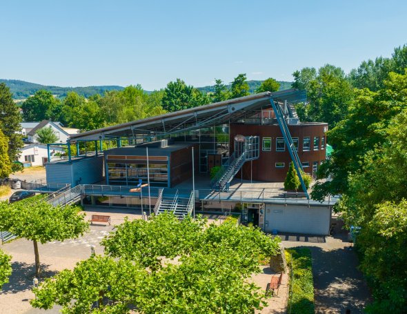 Reichenberghalle Modernes Gebäude mit markantem Schrägdach, großen Glasflächen und Außentreppen, umgeben von Bäumen; davor Platz mit Bänken bei sonnigem Wetter.