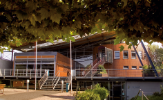 Reichenberghalle Modernes Gebäude mit großem Schrägdach, Holzfassade und Glas, Außentreppen und Geländern, davor Vorplatz mit Bänken und Bäumen bei Sonnenschein.