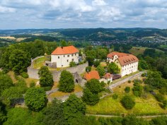 Schloss Reichenberg Luftaufnahme einer Burganlage mit roten Dächern und Ringmauer auf bewaldetem Hügel; dahinter weite Hügellandschaft und Dörfer unter wolkigem Himmel.