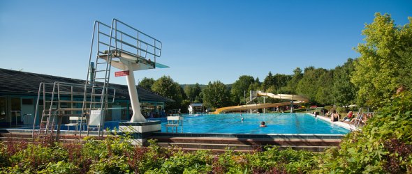 Schwimmbad Reichelsheim Freibad mit Sprungturm, großer blauer Becken, gelber Rutsche und Badenden, umgeben von Bäumen und Beeten unter klarem Sommerhimmel.