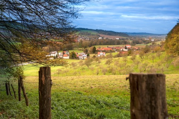 Unter-Gersprenz Blick über grüne Hügel mit Obstbäumen auf ein Dorf mit roten Dächern; Zaunpfähle im Vordergrund, herbstliche Bäume und blauer, bewölkter Himmel.