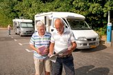 Zwei Reisende lesen Karten vor geparkten Wohnmobilen auf einem Stellplatz am Waldrand; sonniger Tag, weitere Person im Hintergrund.
