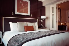 Modernes Hotelzimmer mit großem, gemachtem Bett, Zierkissen und gemusterter Decke; Nachttisch, Lampe und geöffnete Tür zum Bad.