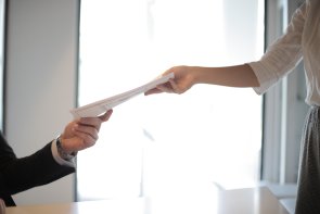 Zwei Personen reichen sich am Schreibtisch ein Dokument; eine Hand nimmt, die andere übergibt, helles Büro im Hintergrund.