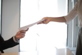 Zwei Personen reichen sich am Schreibtisch ein Dokument; eine Hand nimmt, die andere übergibt, helles Büro im Hintergrund.