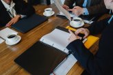 Drei Personen mit Unterlagen Geschäftstreffen am Holztisch: Hände blättern in Ordnern, zeigen auf Unterlagen; Laptops, Notizbücher und Tassen Kaffee stehen bereit.