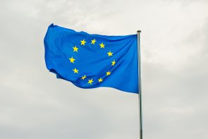 Blaue Europaflagge mit gelben Sternen im Kreis, weht an einem Mast vor grauem, bewölktem Himmel.