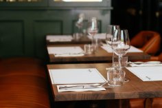 Gedeckter Holztisch im Restaurant: Platzsets, Besteck, Wassergläser und leere Weingläser, daneben Lederbank in warmem Licht.