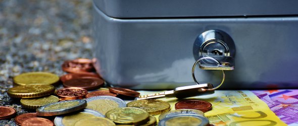 Kleine Geldkassette mit Schlüssel im Schloss; davor liegen verstreute Euromünzen und ein 200-Euro-Schein auf rauer Oberfläche