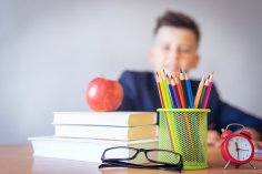 Junge sitzt an Schreibtisch Schreibtisch mit Büchern, Apfel, Stiftebecher, Brille und rotem Wecker; im Hintergrund unscharfe Person. Symbol für Schule oder Lernen.