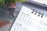 Aufgeschlagener Schreibtischkalender mit Spiralbindung, daneben Stifthalter, bunter Clip und Karotten-Büroklammer auf hellem Untergrund.