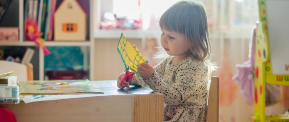Bastelndes Kind Kind sitzt am Tisch in einer Kita und schneidet buntes Papier aus; umgeben von Bastelmaterial, Regal mit Büchern und Spielzeug.
