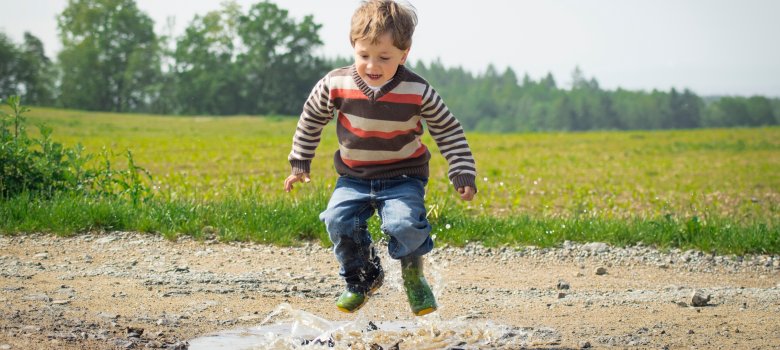 Kind springt in Gummistiefeln in eine Pfütze auf einem Feldweg; Wasser spritzt, im Hintergrund grüne Wiese und Bäume.
