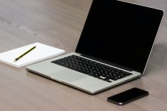 Offenes Laptop auf Holztisch, daneben Notizblock mit Bleistift und ein Smartphone – minimalistischer Arbeitsplatz.