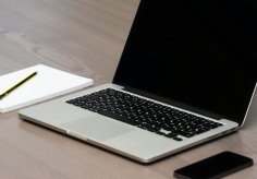 Offenes Laptop auf Holztisch, daneben Notizblock mit Bleistift und ein Smartphone – minimalistischer Arbeitsplatz.