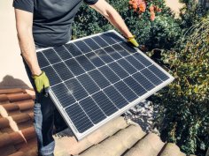 Person mit Handschuhen montiert ein Solarmodul auf einem Ziegeldach, umgeben von Gartenpflanzen.