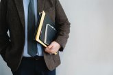 Mann im Anzug Person im Anzug mit braunem Sakko hält Notizbücher und Mappen im Arm, Hand in Hosentasche, neutraler Hintergrund.