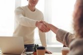 Zwei Personen geben sich am Schreibtisch die Hand, daneben ein Laptop und Kaffeebecher – freundliches Treffen im Büro.