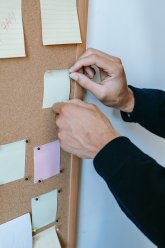 Hände heften einen gelben Haftzettel mit Pinnnadeln an eine Kork-Pinnwand, umgeben von weiteren Notizzetteln in Pastellfarben.