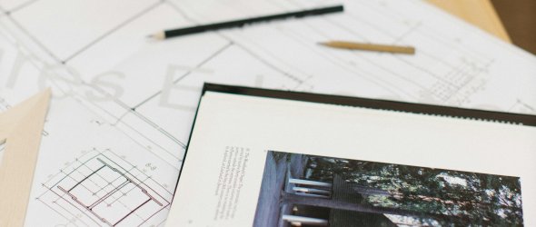 Schreibtisch mit schwarzer Lampe, Brille und Stiften auf Architekturplänen; daneben ein aufgeschlagenes Buch mit Foto eines Gebäudes.