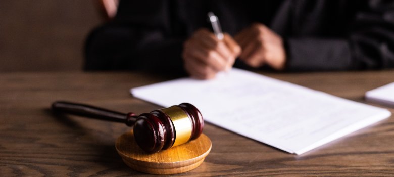 Schreibtisch im Gericht Richterhammer auf Holztisch, im Hintergrund unterschreibt eine Person ein Dokument.